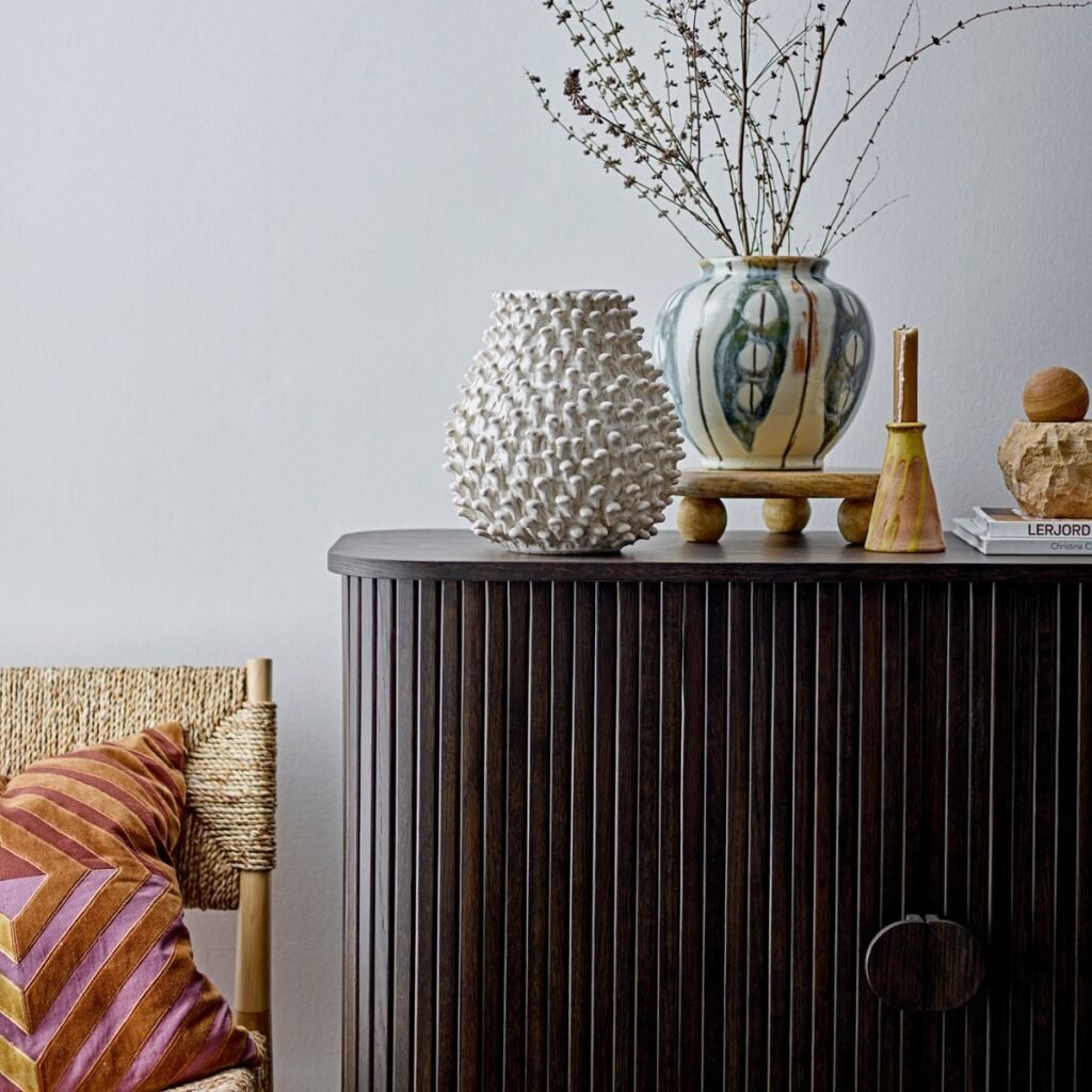 Gros plan sur un buffet en bois foncé avec une finition rainurée, orné de vases en céramique texturée et d'objets décoratifs. À côté, un fauteuil en rotin avec un coussin aux motifs géométriques apporte une touche chaleureuse et naturelle.
