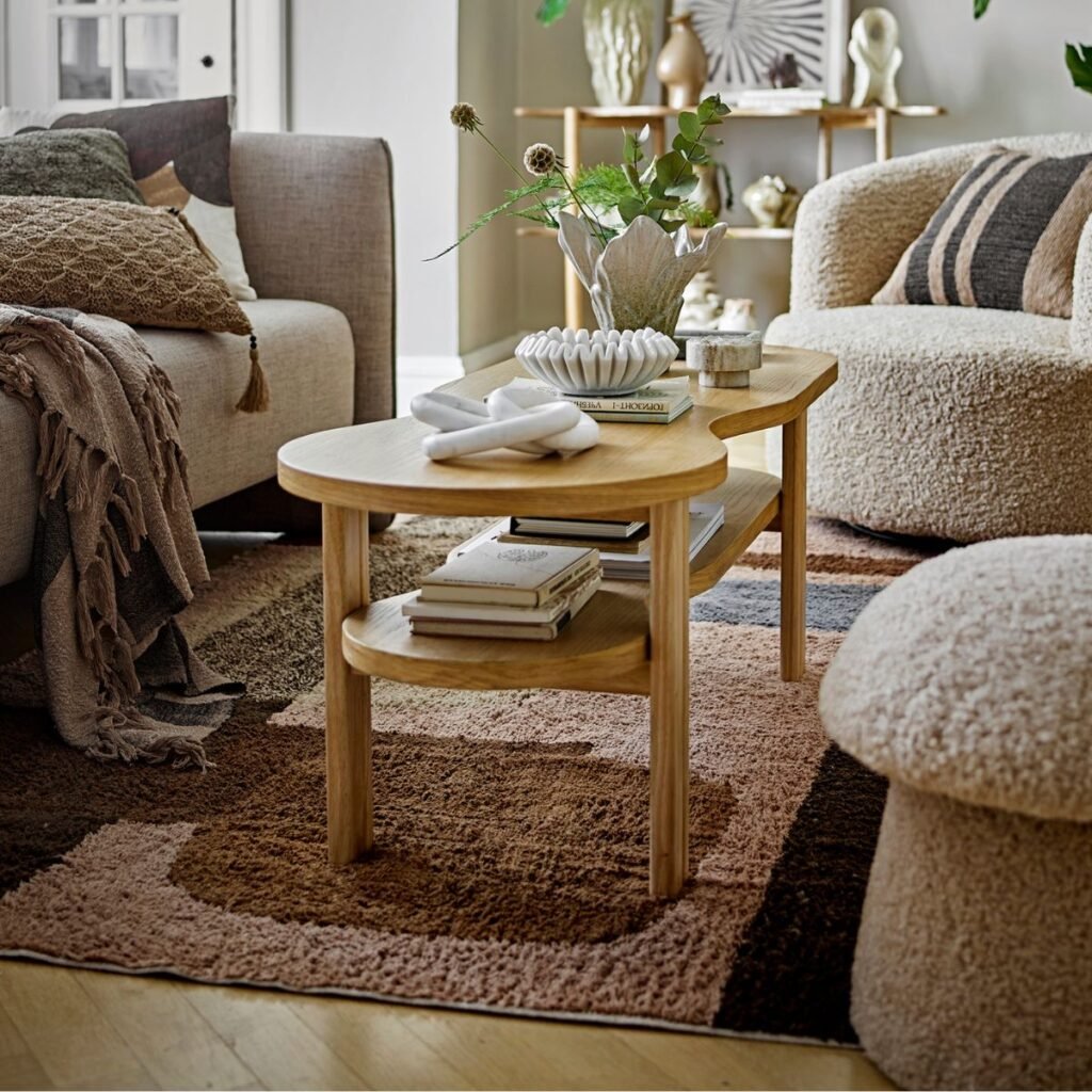 Table basse en bois clair avec une forme organique et une étagère inférieure, placée dans un salon cosy aux tons naturels. Décorée avec des livres, un vase sculptural et des objets en céramique, elle s’intègre harmonieusement à un intérieur chaleureux et texturé.